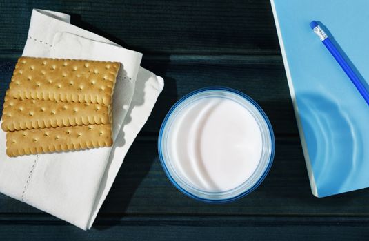 Blue glass of milk on dark blue wooden table , cookies on white napkin and book with pen