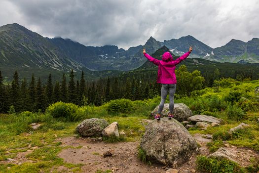 Hooded woman looking at the Tatra mountain panorama in the rain with hands in victory pose - horizontal orientation