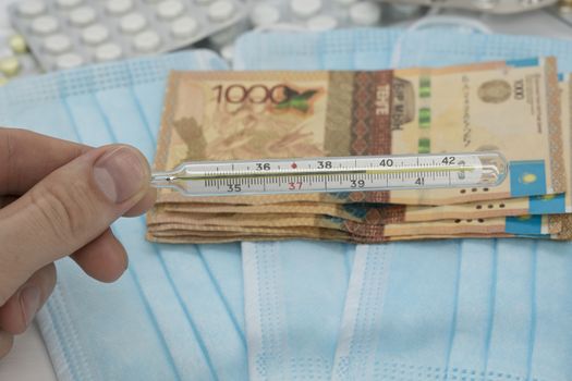 A hand holds a thermometer with a high temperature over a table with pills, a medical mask and tenge money. Increase in the price of medicines in Kazakhstan in tenge KZT. Kazakhstan is quarantined