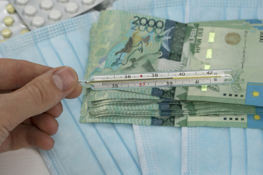 A hand holds a thermometer with a high temperature over a table with pills, a medical mask and tenge money. Increase in the price of medicines in Kazakhstan in tenge KZT. Kazakhstan is quarantined