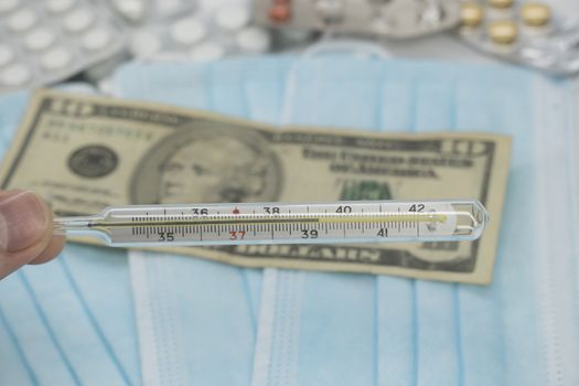 A hand holds a high-temperature thermometer over a table with pills,a medical mask and dollar money.The growth of prices for medicines in America in dollars.The United States is under quarantine