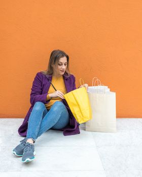 A middle-aged blonde woman sitting on the floor looking at shopping bags on an orange background