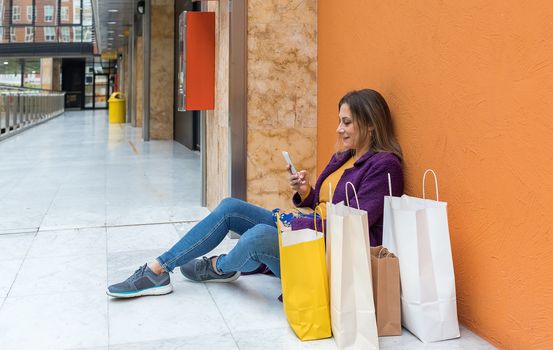 Adult woman sitting on the floor of a shopping mall looking at the mobile phone with shopping bags next to her