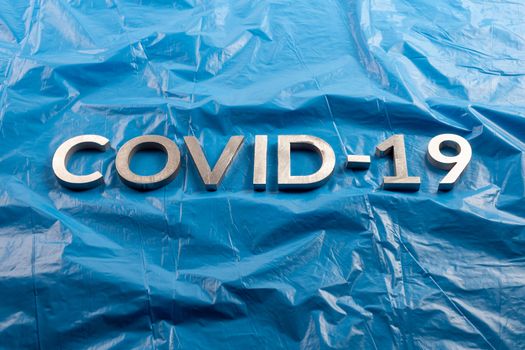 The word COVID-19 laid with aluminium letters on crumpled blue plastic film background in flat lay composition.