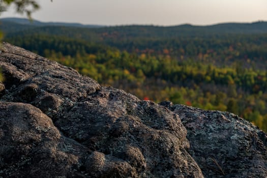 The view of the edge of a cliff in autumn shows the detailed texture of its rocky surface before a blurred background of autumn forest trees.