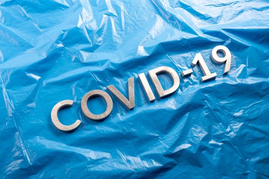 The word COVID-19 laid with aluminium letters on crumpled blue plastic film background in flat lay composition, diagonal perspective.