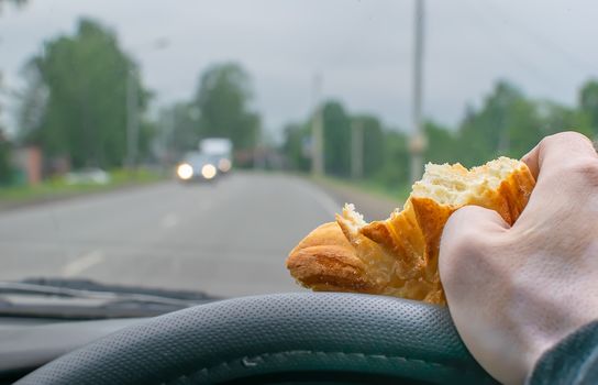 a bitten bun in the hand of a car driver who is holding the steering wheel while on the roadway in a locality