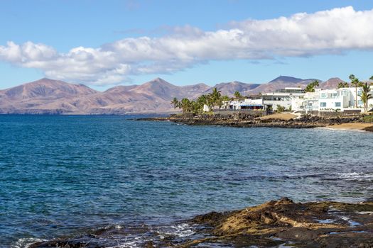 Rocky coast of Puerto del Carmen at Canary island Lanzarote with lava rocks and blue water in the foreground, white houses, palm trees and volcanic mountain range with different colours the background