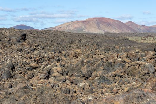 Volcanic landscape in the south west of canary island Lanzarote with dark lava rocks in the foreground and red volcanic mountains in the background. The sky is blue with clouds. 