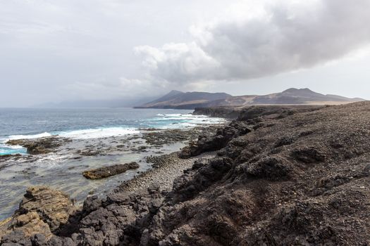 Panoramic view at the coastline in the natural park of Jandia (Parque Natural De Jandina) on canary island Fuerteventura, Spain with gravel, lava rocks and rough sea with waves and mountain range