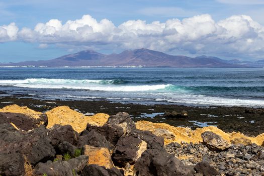 Panoramic view at the coastline of Corralejo on canary island Fuerteventura, Spain with  lava rocks and rough sea with waves and canary island Lanzarote in the  background