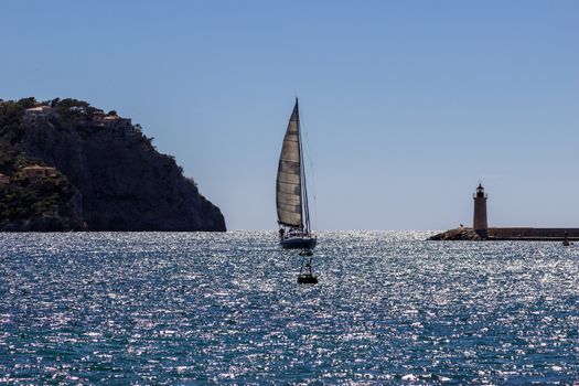  Sailboat in the harbor entrance of Port d' Andratx between a Lighthouse and a mountain with houses