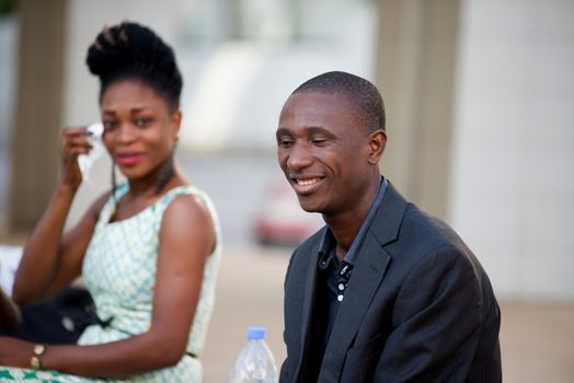 smiling young businessman sitting next to a young woman looking at him and wiping his face with a white pocket in his hand.
