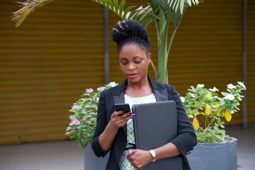 young businesswoman standing with a laptop, reading messages in her mobile phone.