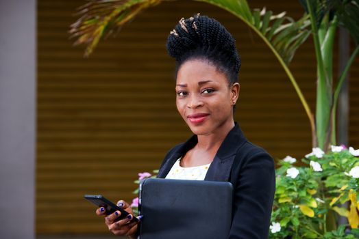 Smiling businesswoman using smart phone while standing outside holding a laptop.