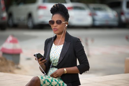 young woman wearing a black jacket and sunglasses sits outside and listens to music with her mobile phone