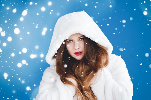 Happy Christmas and winter holiday portrait of young woman in white hooded fur coat, snow on blue background, fashion and lifestyle campaign