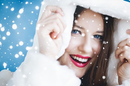 Happy Christmas and winter holiday portrait of young woman in white hooded fur coat, snow on blue background, fashion and lifestyle campaign