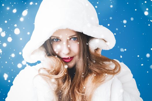 Happy Christmas and winter holiday portrait of young woman in white hooded fur coat, snow on blue background, fashion and lifestyle campaign