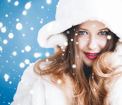 Happy Christmas and winter holiday portrait of young woman in white hooded fur coat, snow on blue background, fashion and lifestyle campaign