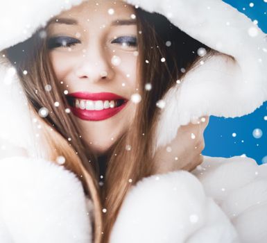 Happy Christmas and winter holiday portrait of young woman in white hooded fur coat, snow on blue background, fashion and lifestyle campaign