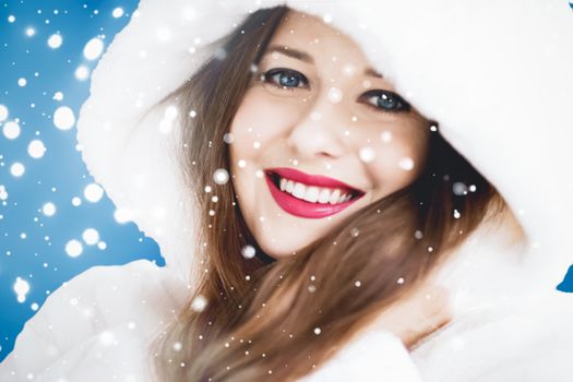 Happy Christmas and winter holiday portrait of young woman in white hooded fur coat, snow on blue background, fashion and lifestyle campaign