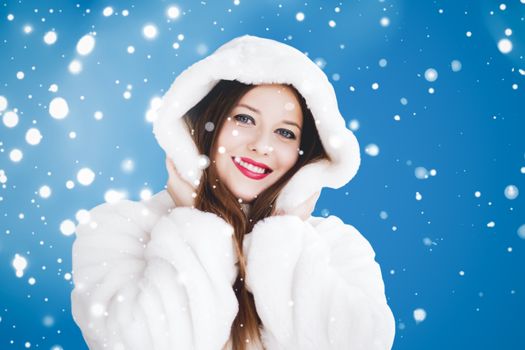 Happy Christmas and winter holiday portrait of young woman in white hooded fur coat, snow on blue background, fashion and lifestyle campaign