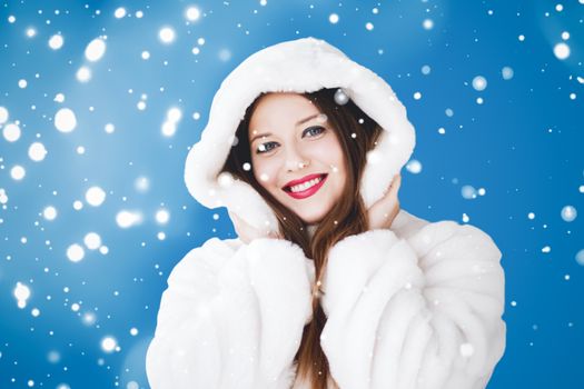 Happy Christmas and winter holiday portrait of young woman in white hooded fur coat, snow on blue background, fashion and lifestyle campaign