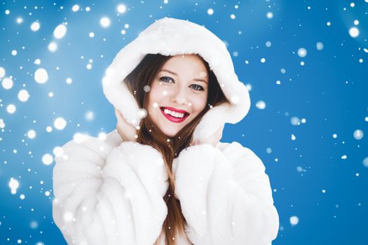 Happy Christmas and winter holiday portrait of young woman in white hooded fur coat, snow on blue background, fashion and lifestyle campaign