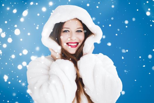 Happy Christmas and winter holiday portrait of young woman in white hooded fur coat, snow on blue background, fashion and lifestyle campaign