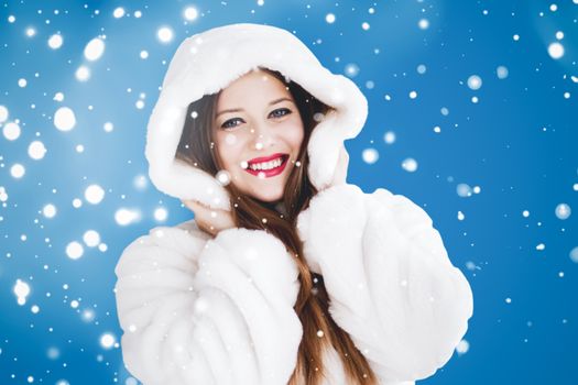 Happy Christmas and winter holiday portrait of young woman in white hooded fur coat, snow on blue background, fashion and lifestyle campaign