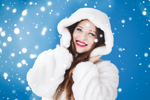 Happy Christmas and winter holiday portrait of young woman in white hooded fur coat, snow on blue background, fashion and lifestyle campaign