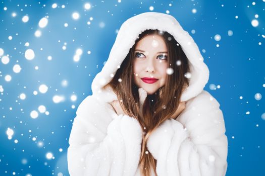 Happy Christmas and winter holiday portrait of young woman in white hooded fur coat, snow on blue background, fashion and lifestyle campaign