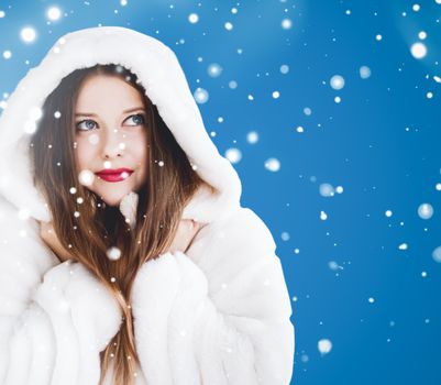 Happy Christmas and winter holiday portrait of young woman in white hooded fur coat, snow on blue background, fashion and lifestyle campaign