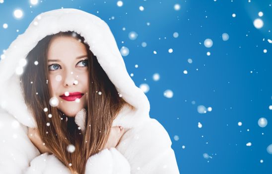 Happy Christmas and winter holiday portrait of young woman in white hooded fur coat, snow on blue background, fashion and lifestyle campaign