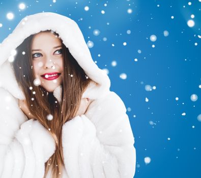 Happy Christmas and winter holiday portrait of young woman in white hooded fur coat, snow on blue background, fashion and lifestyle campaign