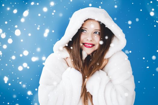 Happy Christmas and winter holiday portrait of young woman in white hooded fur coat, snow on blue background, fashion and lifestyle campaign