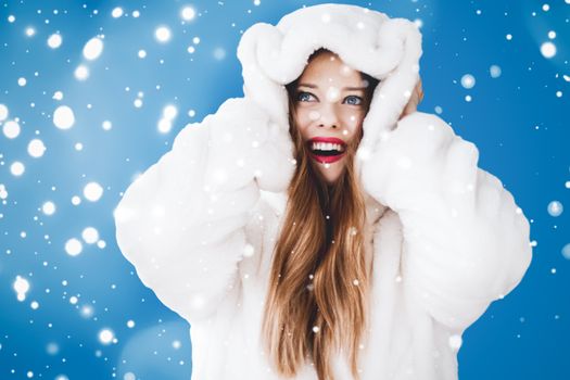 Happy Christmas and winter holiday portrait of young woman in white hooded fur coat, snow on blue background, fashion and lifestyle campaign