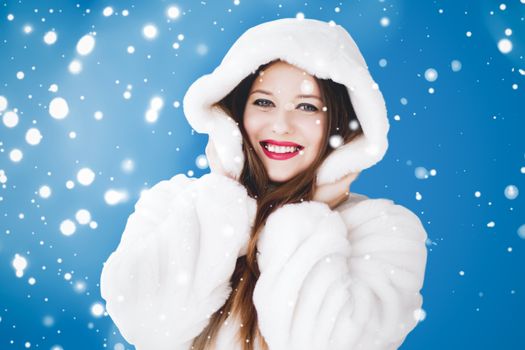 Happy Christmas and winter holiday portrait of young woman in white hooded fur coat, snow on blue background, fashion and lifestyle campaign