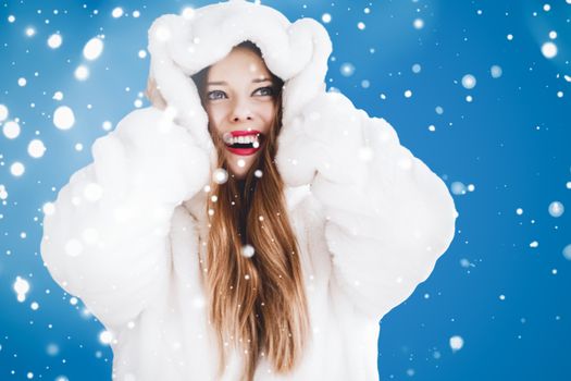 Happy Christmas and winter holiday portrait of young woman in white hooded fur coat, snow on blue background, fashion and lifestyle campaign