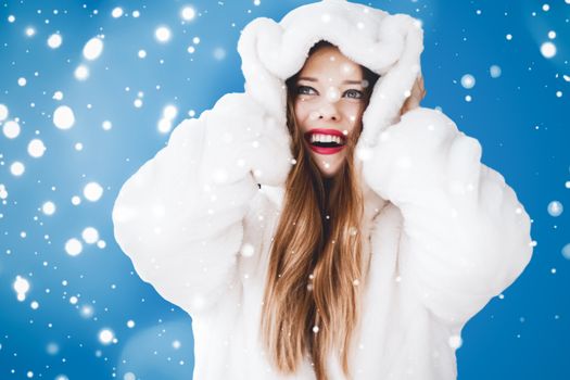 Happy Christmas and winter holiday portrait of young woman in white hooded fur coat, snow on blue background, fashion and lifestyle campaign