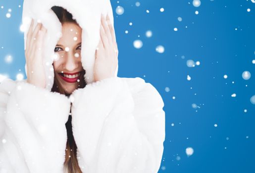 Happy Christmas and winter holiday portrait of young woman in white hooded fur coat, snow on blue background, fashion and lifestyle campaign