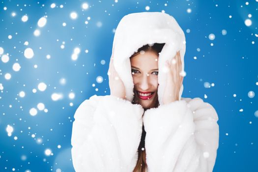 Happy Christmas and winter holiday portrait of young woman in white hooded fur coat, snow on blue background, fashion and lifestyle campaign