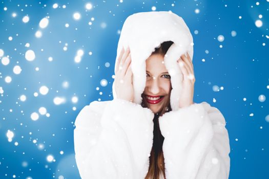 Happy Christmas and winter holiday portrait of young woman in white hooded fur coat, snow on blue background, fashion and lifestyle campaign