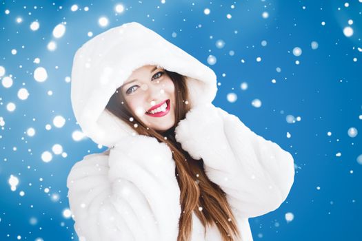 Happy Christmas and winter holiday portrait of young woman in white hooded fur coat, snow on blue background, fashion and lifestyle campaign