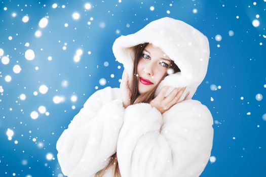 Happy Christmas and winter holiday portrait of young woman in white hooded fur coat, snow on blue background, fashion and lifestyle campaign