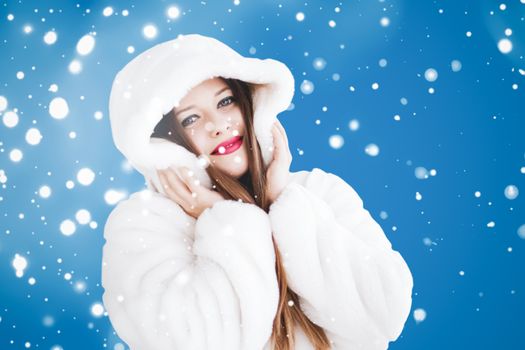 Happy Christmas and winter holiday portrait of young woman in white hooded fur coat, snow on blue background, fashion and lifestyle campaign