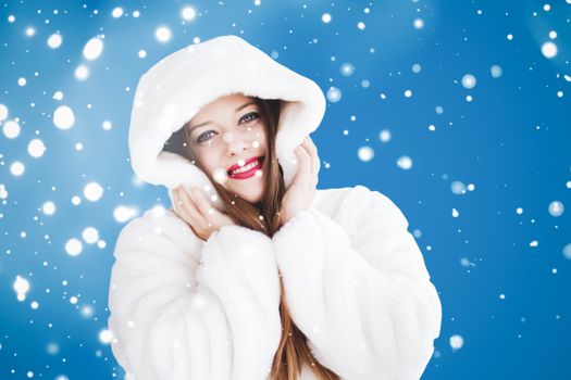 Happy Christmas and winter holiday portrait of young woman in white hooded fur coat, snow on blue background, fashion and lifestyle campaign