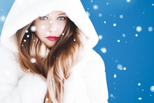 Happy Christmas and winter holiday portrait of young woman in white hooded fur coat, snow on blue background, fashion and lifestyle campaign