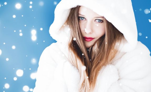 Happy Christmas and winter holiday portrait of young woman in white hooded fur coat, snow on blue background, fashion and lifestyle campaign